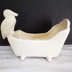 Pode incluir: Um vaso de cerâmica branco em forma de banheira, com um pássaro empoleirado na lateral. O vaso tem três pés pequenos e um interior curvo, adequado para pequenas plantas ou itens decorativos.