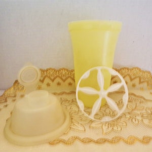 Puede incluir: Un juego de tres utensilios de cocina de plástico amarillo, que incluye una taza, una tapa y una herramienta en forma de estrella. Los utensilios están sobre un mantel de encaje.