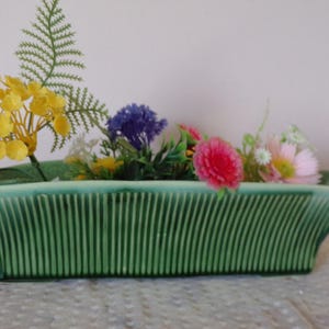 May include: A rectangular green ceramic planter with a vertical ribbed texture on the exterior sides. The planter is filled with an assortment of artificial flowers, including yellow, purple, pink, and white blossoms along with green fern-like foliage.