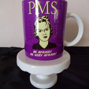 Könnte beinhalten: Eine lila Keramiktasse mit dem Wort "PMS" in großen, cremefarbenen Buchstaben. Darunter eine Vintage-Illustration einer Frau mit wütendem Gesichtsausdruck. Die Tasse trägt auch den Text "BE AFRAID! BE VERY AFRAID!" und einen weißen Henkel.