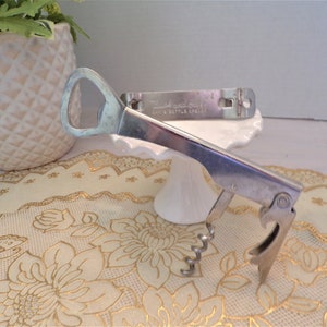 Puede incluir: Un abridor de botellas y sacacorchos combinado plateado vintage. La herramienta tiene un abridor de botellas, un sacacorchos y un abrelatas. El texto "Hand and Seal CAN & BOTTLE OPENER" es visible en un abrelatas separado. Los artículos están sobre una superficie blanca.