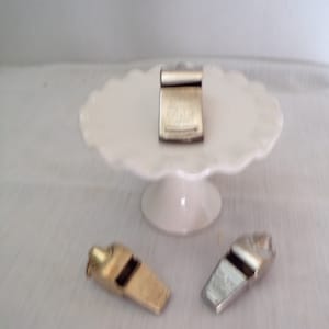 May include: Three whistles, one silver and two gold, on a white cake stand. The silver whistle has a black and white design on the side. The gold whistles have a smooth surface.