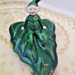 May include: A green ceramic figurine of a gnome sitting on a large green leaf. The gnome is wearing a green hat and a green outfit.