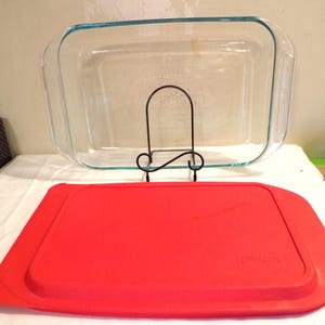May include: A clear rectangular glass baking dish with a blue rim, resting on a black metal stand, and a bright red lid. The lid is rectangular and covers the dish. The dish is designed for baking and food storage.