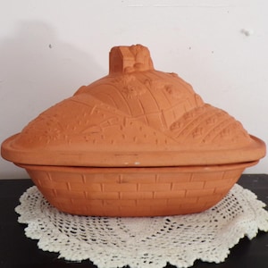 May include: An oval terracotta baking dish with a lid featuring a raised relief design of a landscape with a small house. The base of the dish has a textured brick pattern, and the lid is topped with a small handle shaped like a building.