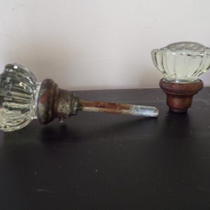 Può includere: Due pomelli per porta con impugnature in vetro trasparente scanalato fissate su basi in metallo scuro e usurato. Un pomello è posizionato orizzontalmente con un lungo perno metallico arrugginito, mentre l'altro è in posizione verticale.
