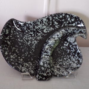 May include: A black glass dish with an irregular, organic shape featuring a speckled white pattern throughout the surface. The dish has a raised, curved ridge that divides the interior space and is supported by two small, clear plastic stands.