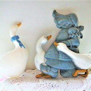 May include: A blue ceramic figurine of a girl holding two white ceramic geese. The girl is wearing a blue dress and a white apron. The geese have blue ribbons around their necks.