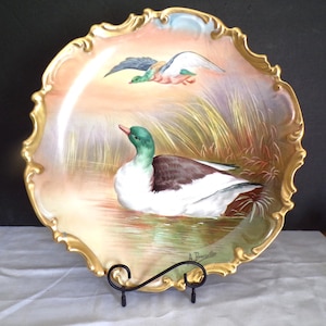 May include: Ornate porcelain plate with a hand-painted duck scene. The plate has a gold, scalloped edge and depicts ducks in a marsh setting. The plate is displayed on a black metal stand. The artwork includes ducks in flight and on the water.