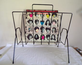 May include: A black metal magazine rack holding a vintage Rolling Stones album. The album cover features multiple portraits of women with colorful accents. The rack has a rectangular shape with a handle at the top and angled legs.