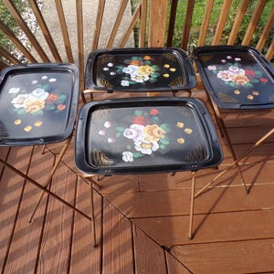May include: A set of four black metal TV tray tables with folding legs. Each rectangular tray features a hand-painted floral design with white, yellow, and red roses, green leaves, and gold-toned decorative line accents on the corners.