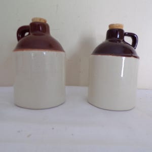 Puede incluir: Dos jarras de cerámica con tapa y asa marrones y cuerpo beige. Cada jarra tiene un tapón de corcho. Las jarras son probablemente para fines decorativos o para almacenar líquidos. Las jarras están sobre una superficie blanca.