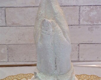 May include: A white, glitter-covered sculpture of hands in prayer. The hands are clasped, fingers pointing upwards. The sculpture sits on a white, scalloped plate, against a brick wall.