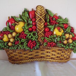 May include: A wall-mounted decorative plaque shaped like a woven basket filled with a bounty of fruit. The relief features red apples, yellow pears, and clusters of red berries nestled among green leaves, all set against a brown wicker-textured base.