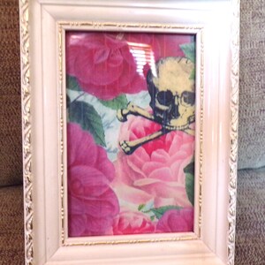 Könnte beinhalten: Ein weißer Bilderrahmen mit goldener Verzierung, der ein Totenkopf- und Knochenkreuzmotiv auf einem Hintergrund aus rosa Rosen zeigt.