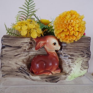 May include: A ceramic planter shaped like a log, featuring a brown deer figurine and artificial yellow flowers. The planter has a textured, wood-grain appearance and a light green leaf detail.