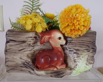 May include: A ceramic planter shaped like a log, featuring a brown deer figurine and artificial yellow flowers. The planter has a textured, wood-grain appearance and a light green leaf detail.