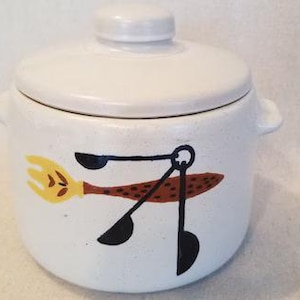 May include: A white ceramic cookie jar with a lid and two small handles. The jar features a whimsical design of a yellow whisk, a brown fish, and black utensils. The jar is a kitchenware item.