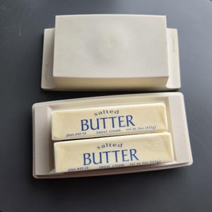 May include: A light gray butter dish with a lid, holding two sticks of salted butter. The butter is labeled "BUTTER" with "sweet cream" and "net wt 4oz (113g)" printed on the wrapper. The dish is on a dark surface.