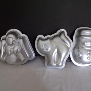 May include: Three metal baking molds in the shapes of an angel, a cat, and a snowman. The molds are silver-colored and have a simple, outlined design. The angel has wings, the cat is standing, and the snowman has a hat and buttons.
