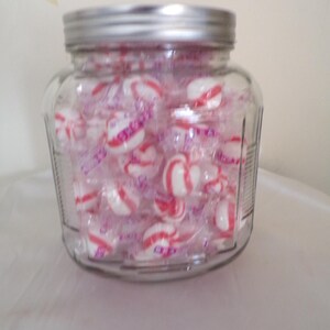 Puede incluir: Un tarro de cristal transparente lleno de caramelos envueltos individualmente. Los caramelos son blancos con rayas rojas y están envueltos en celofán transparente. El tarro tiene una tapa de metal plateado.