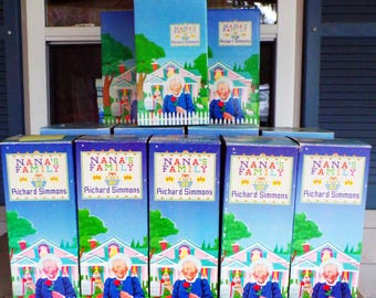 Può includere: Una collezione di scatole blu con la scritta "NANA'S FAMILY" e "Richard Simmons". Ogni scatola mostra un'illustrazione di una donna in abito blu che tiene una rosa davanti a una casa con una staccionata bianca. Le scatole sono impilate.