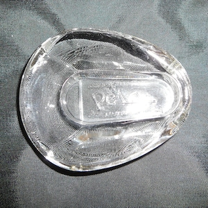 May include: A clear glass ashtray with a raised oval in the center. The ashtray has a "D'Arcy" logo etched into the bottom.
