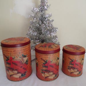Pode incluir: Conjunto de três latas redondas de metal com tampas. As latas apresentam um tema musical com um violino vermelho, partituras e azevinho sobre um fundo laranja. As tampas e as bordas são de cor vermelho escuro. Uma árvore de Natal prateada de oropel está no fundo.