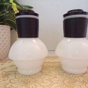 Puede incluir: Dos botellas de vidrio blanco con tapas de plástico marrón. Las botellas tienen un fondo redondeado y una parte superior ancha y plana. Las botellas están sobre un mantel de encaje.