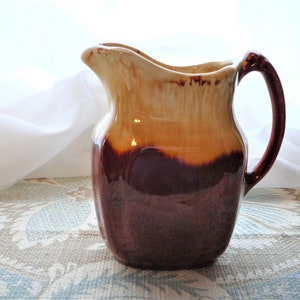 May include: A brown and cream glazed ceramic pitcher with a handle. The pitcher has a rustic, vintage look.