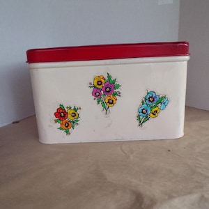 Könnte beinhalten: Rechteckige weiße Metall-Aufbewahrungsbox mit rotem Deckel. Die Box ist mit drei Clustern farbenfroher Blumendesigns verziert. Die Blumen umfassen gelbe, violette, rote und blaue Blüten. Wahrscheinlich Vintage.