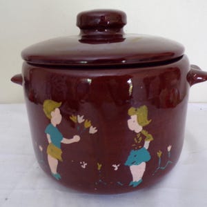 Może przedstawiać: Brązowy ceramiczny słoik na ciastka z pokrywką i bocznymi uchwytami. Słoik jest ozdobiony malowanymi postaciami dzieci i kwiatów. Słoik jest prawdopodobnie w stylu vintage i nadaje się do dekoracji kuchni.