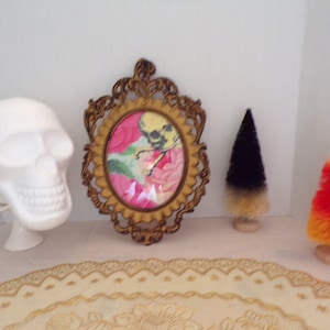 May include: A white ceramic skull figurine, a gold ornate frame with a floral print and a skull image, and two small brush trees, one black and one orange and yellow, on a white surface.