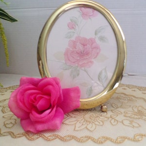 May include: An oval gold-colored picture frame with a floral print featuring pink roses. A vibrant pink rose rests on a cream-colored lace surface, complementing the frame's design. The frame is supported by small gold feet.