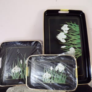 May include: A set of three black rectangular serving trays with gold-toned edges. Each tray features a graphic illustration of white iris flowers with green stems and leaves. Two of the trays are partially covered in clear plastic wrap.