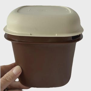 May include: A brown and beige plastic food container with a lid. The container is held by a hand. The container is square with rounded edges. The lid is beige and fits snugly on top. The container is brown.
