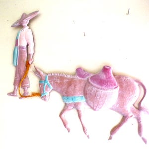 May include: A pink and white figurine of a person wearing a sombrero and a long, flowing robe, leading a pink donkey carrying a large, pink, woven basket.