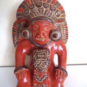 May include: A red ceramic figurine of a figure with a headdress and ear ornaments. The figure has a detailed face and is wearing a decorative collar with the word "eco". The figurine is a unique piece of art.