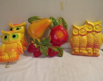 May include: A collection of vintage wall decor featuring three owl figurines and a fruit-themed piece. The owls are primarily yellow with orange accents, while the fruit includes a pear, apple, and strawberries. The pieces are made of a glossy material.