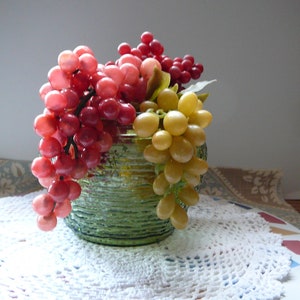May include: A green glass vase with a textured surface holds a cluster of red and yellow grapes. The vase is sitting on a white crocheted doily.