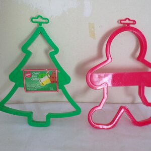 Puede incluir: Dos cortadores de galletas de plástico: un árbol de Navidad verde y un hombre de jengibre rojo. El cortador de árbol tiene una etiqueta que dice "Giant Tree Cutter". Ambos cortadores tienen un lazo para colgar.