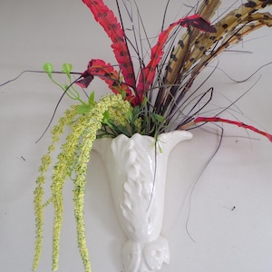 Puede incluir: Un jarrón de pared de cerámica blanca con un diseño floral decorativo, que contiene un ramo de flores artificiales rojas, marrones y amarillas.