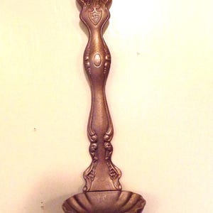 May include: Ornate bronze-colored spoon with a decorative handle and a small, scalloped bowl. The handle features detailed scrollwork and a shield-like emblem. A vintage-style utensil.