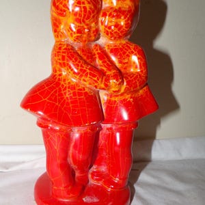 May include: A vintage ceramic figurine of two figures embracing, finished in a glossy red glaze with a cracked, textured pattern. The figures are dressed in matching outfits, standing on a round base.