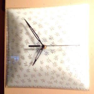 May include: A white square clock with black hands and a silver starburst pattern. The clock is framed in a clear glass frame.