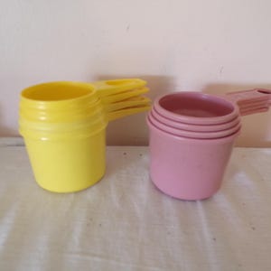 May include: Two sets of nested plastic measuring cups, one set in bright yellow and the other in a muted pink shade. Each set features multiple cups stacked together with integrated handles extending from the side of each individual cup unit.