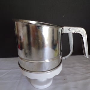 May include: A vintage metal flour sifter with a handle and a white ceramic base. The sifter is made of shiny, reflective metal and has a handle for easy use. The white base has a decorative, scalloped edge.