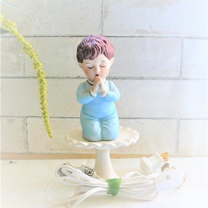May include: A porcelain figurine of a boy with brown hair, wearing a blue outfit, praying with his hands clasped together. The figurine is on a white pedestal base and is illuminated by a light bulb. The figurine is connected to a white electrical cord.