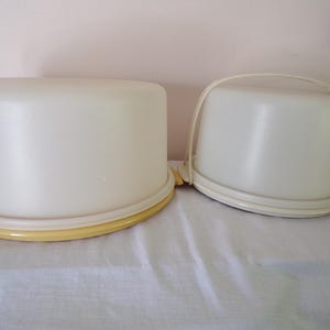 Puede incluir: Dos porta tartas de plástico blanquecino. El más grande es redondo con base amarilla. El más pequeño tiene asa y base blanca. Ambos tienen tapa abovedada. Están sobre una superficie blanca.
