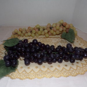 May include: Two bunches of artificial grapes, one dark purple and the other a mix of green and pink, sit on a decorative, gold-edged doily. Green leaves are attached to the purple grapes. The grapes are a decorative item.